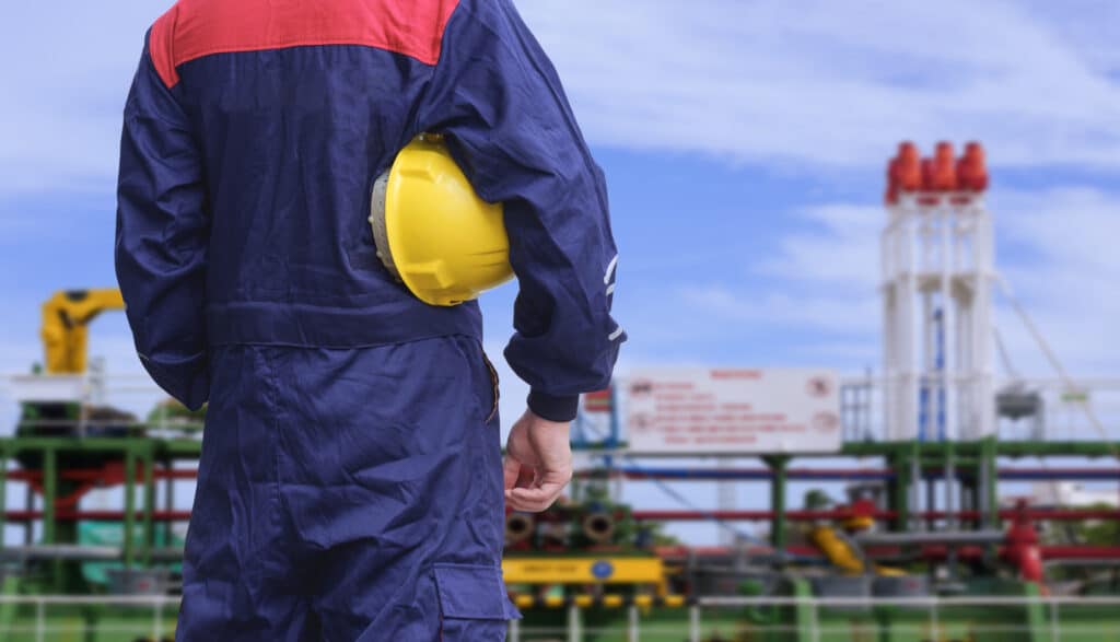 Offshore worker stands looking out at oil tanker.